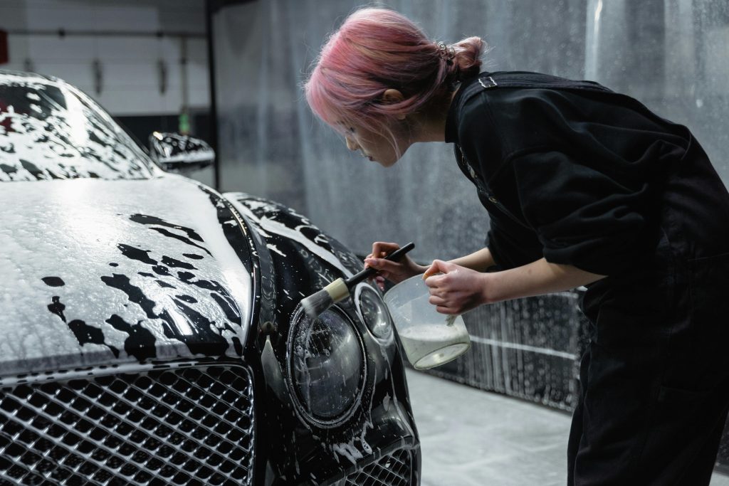 A woman with pink hair meticulously cleans a black car's headlight at an indoor car wash.