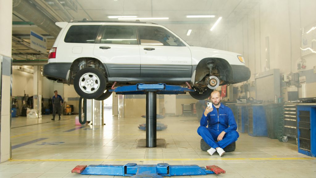 Mechanic in blue coverall using smartphone beside vehicle on lift in repair workshop.